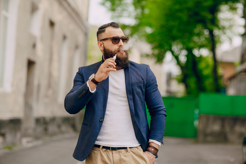 Un homme barbu en train de fumer une cigarette électronique expire en marchant dans la rue.