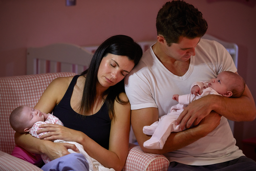Une femme tenant un bébé a les yeux fermés et est appuyée contre un homme qui tient aussi un bébé; les deux sont assis sur un lit.