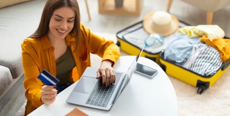 A person shops online with a credit card beside an open suitcase filled with travel items, suggesting booking eligible travel purchases such as flights, hotels, car rentals, and vacation cruises.