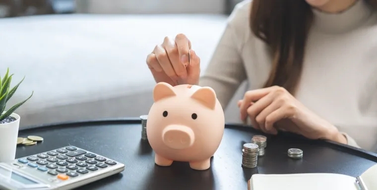 A person drops a coin into a piggy bank on a table surrounded by coins, a calculator, and a notebook, symbolizing saving money and redeeming points for gift cards.