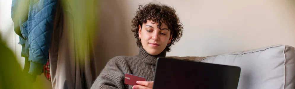 A woman sits on a sofa with a laptop, looking at a credit card in her hand, considering her online payment options.