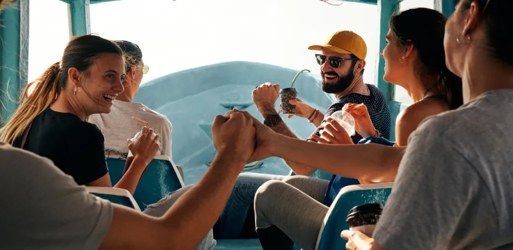Un groupe de personnes voyage ensemble dans un petit bateau ouvert, se tenant les mains en cercle et profitant du moment tandis que la lumière du soleil éclaire l’avant du bateau sur l’eau.