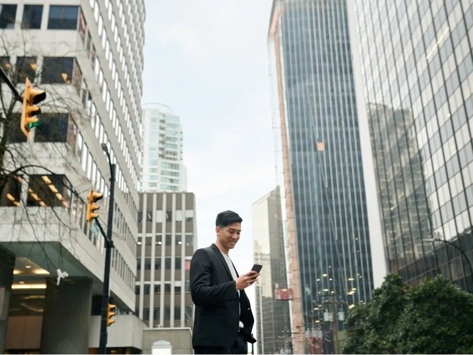 A person stands on a city street surrounded by tall office buildings, looking at their phone while traffic lights and urban architecture frame the scene.