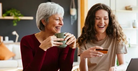 A mother smiles at her daughter holding a credit card as they view a laptop screen.